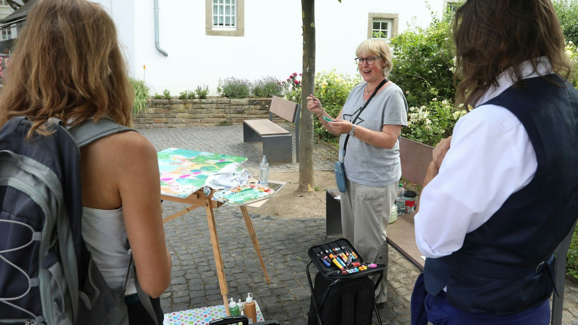 Eine Künstlerin hat eine Staffelei aufgebaut und spricht mit zwei Besuchern.