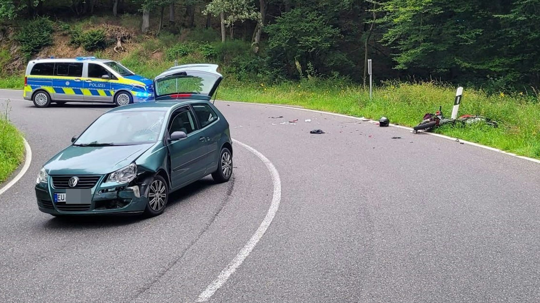 Auf der linken Seite ist ein beschädigter Wagen zu sehen, am Straßenrand ein auf der Erde liegendes Motorrad. Im Hintergrund steht ein Polizeiwagen.Teile der verunfallten Fahrzeuge liegen auf der Straße.