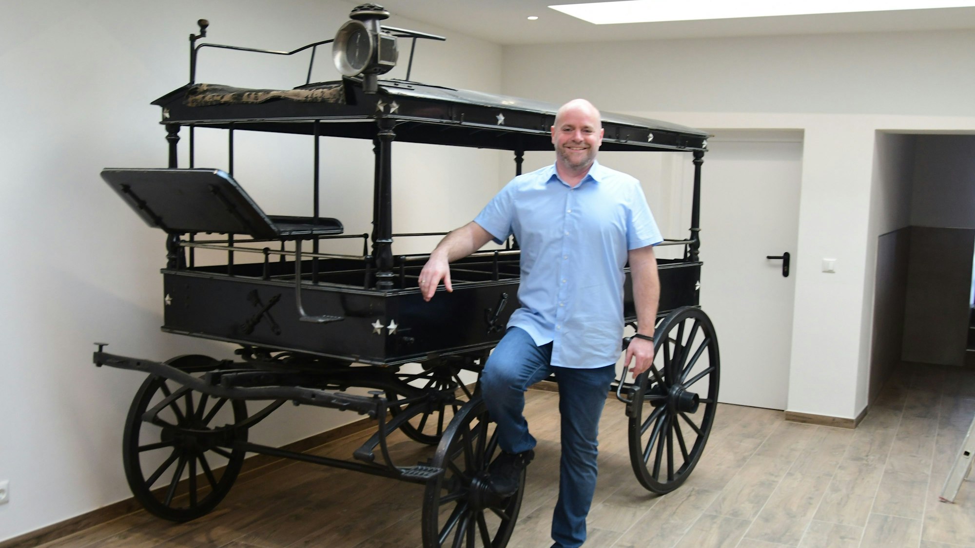 Bestatter Gerd Lethert aus Bad Münstereifel steht in seinen frisch sanierten Räumen an einer historischen, schwarzen Kutsche.