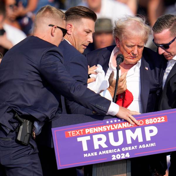 Sicherheitsbeamte schirmen den amerikanischen Ex-Präsidenten Donald Trump ab. er hat Blut im Gesicht, seine Haare sind durcheinander.