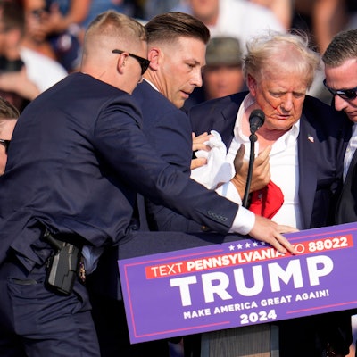 Sicherheitsbeamte schirmen den amerikanischen Ex-Präsidenten Donald Trump ab. er hat Blut im Gesicht, seine Haare sind durcheinander.