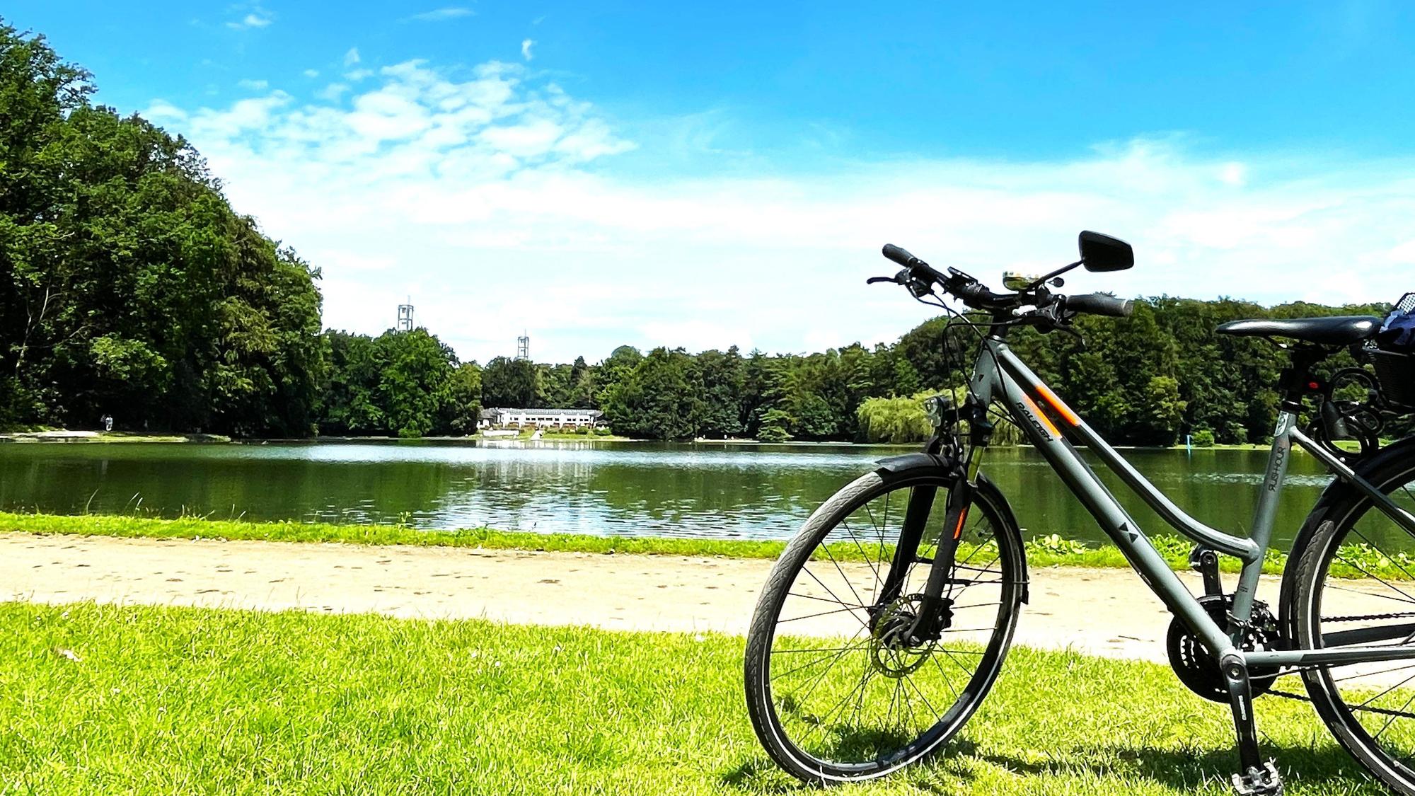 Ein Fahrrad steht auf der Wiese am Adenauerweiher in Köln. In der Ferne sind der Club Astoria und die Stützen vom Rhein Energie Stadion zu sehen.