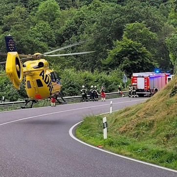 Wenige Meter über der Straße ist gelber ADAC-Rettungshubschrauber zu sehen. Im Hintergrund stehen Einsatzkräfte und ein Feuerwehrwagen.