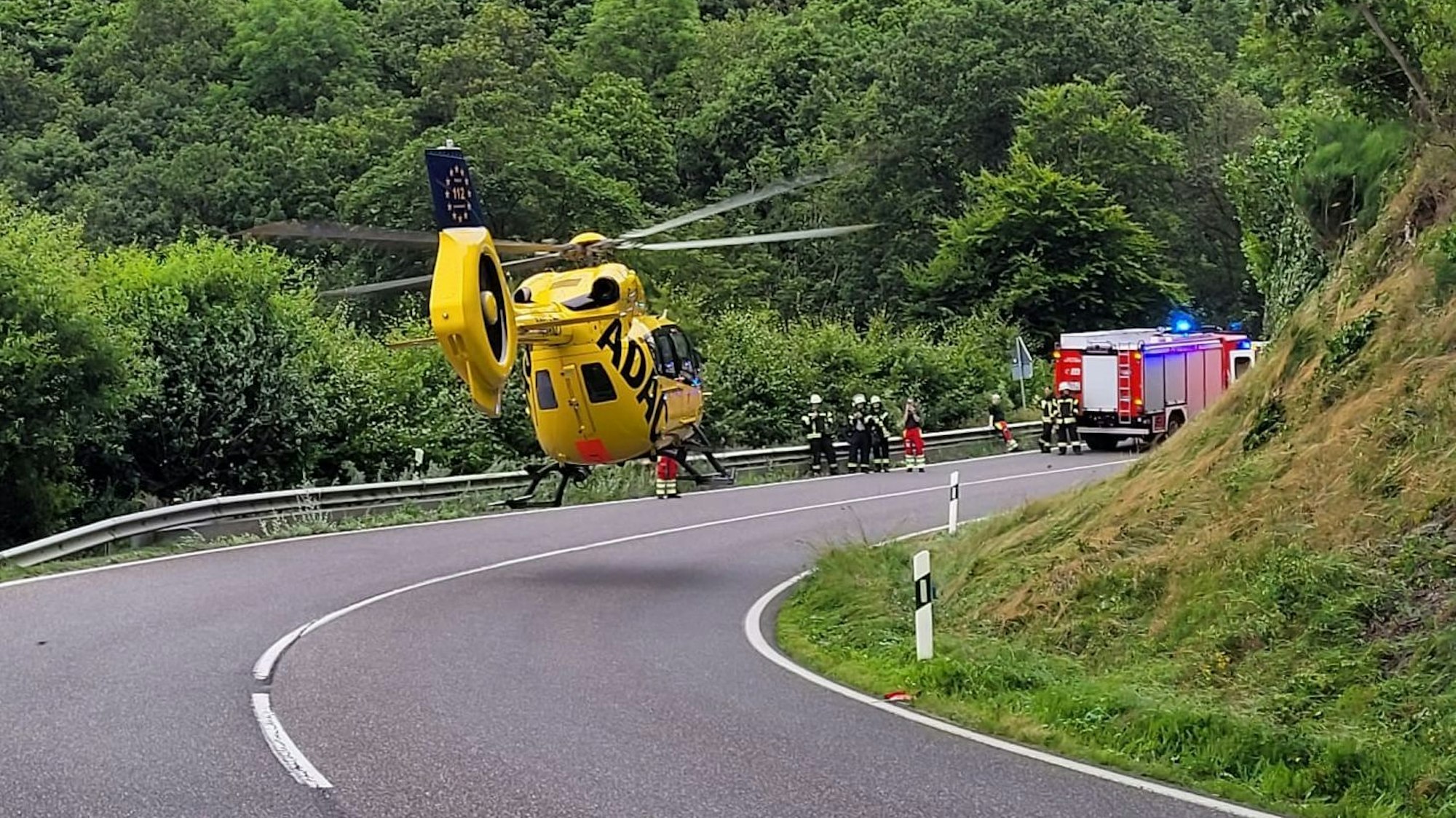 Wenige Meter über der Straße ist gelber ADAC-Rettungshubschrauber zu sehen. Im Hintergrund stehen Einsatzkräfte und ein Feuerwehrwagen.
