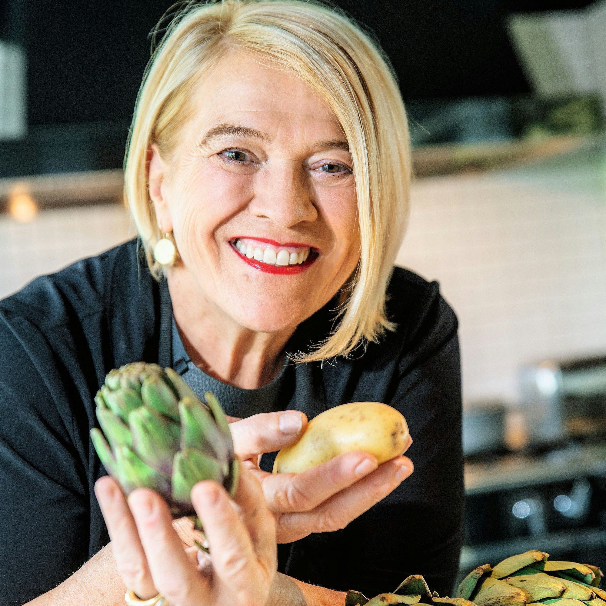 Lea Linster hält eine Artischocke in der einen und eine Kartoffel in der anderen Hand. Sie trägt eine schwarze Kochjacke.
