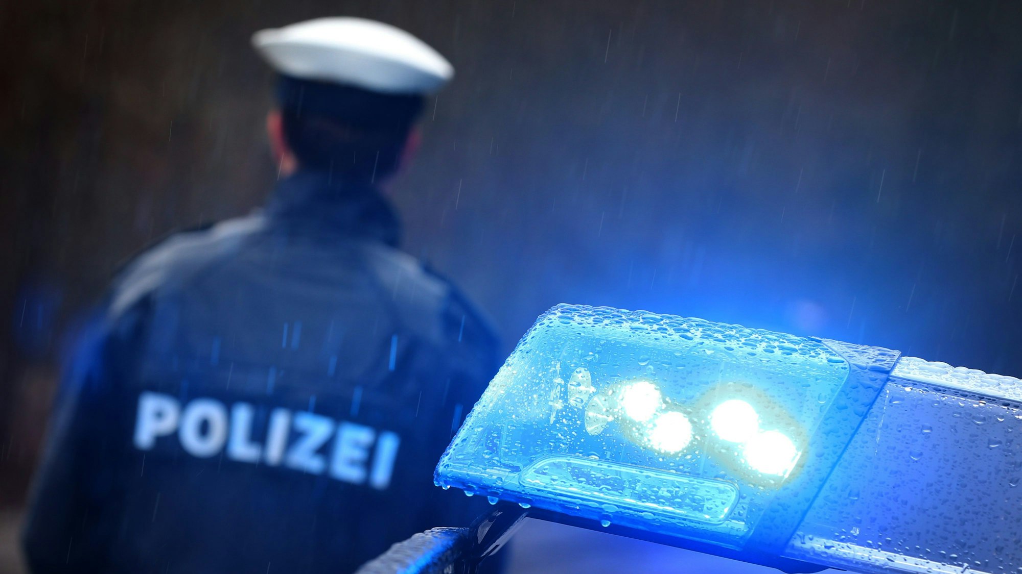 Ein Polizist steht im Regen vor einem Streifenwagen dessen Blaulicht aktiviert ist. (Symbolbild)
