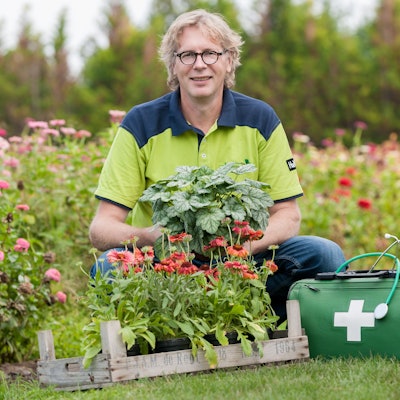 René Wadas posiert neben einem Arztkoffer inmitten blühender Blumen.