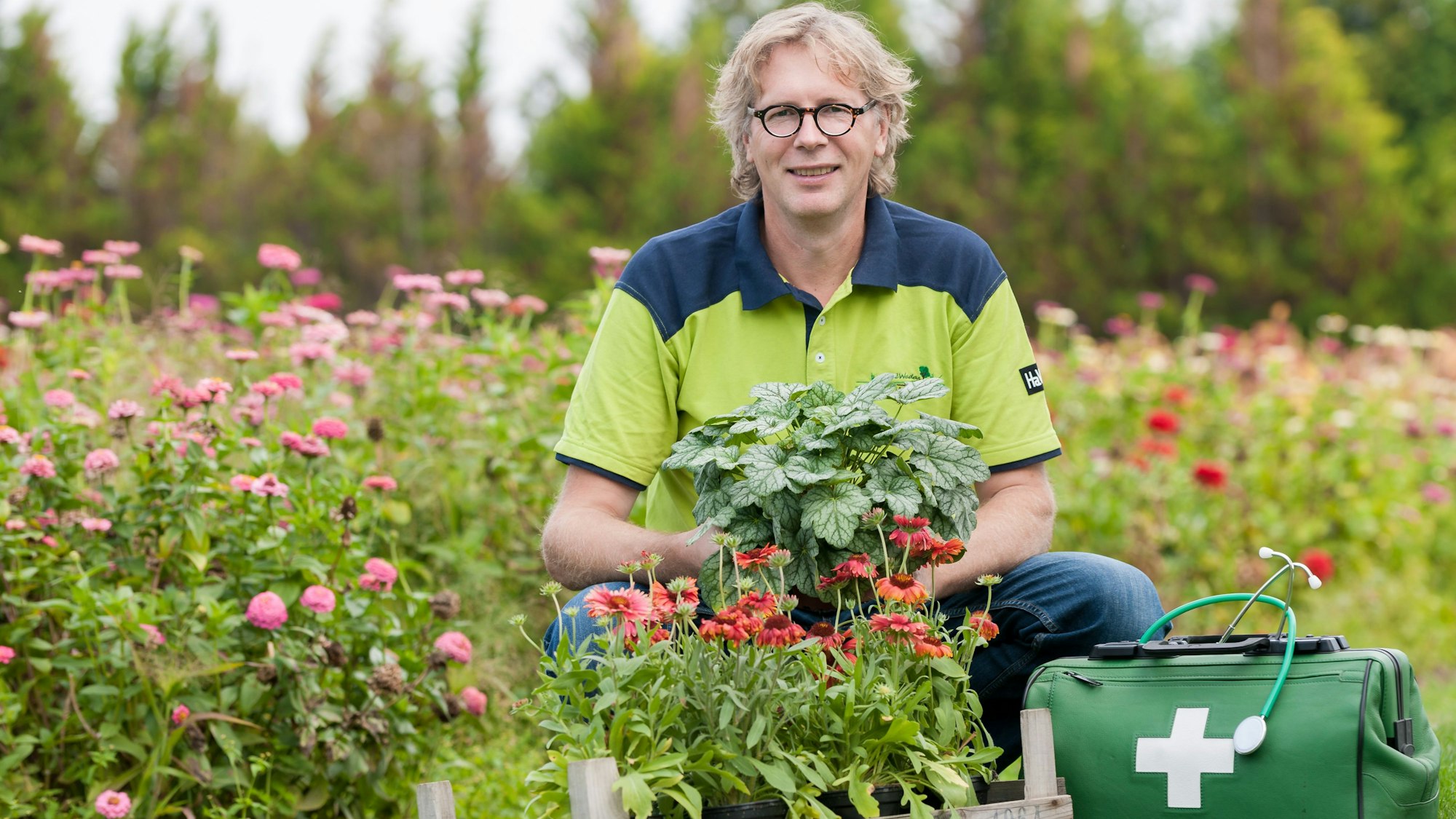 René Wadas posiert neben einem Arztkoffer inmitten blühender Blumen.