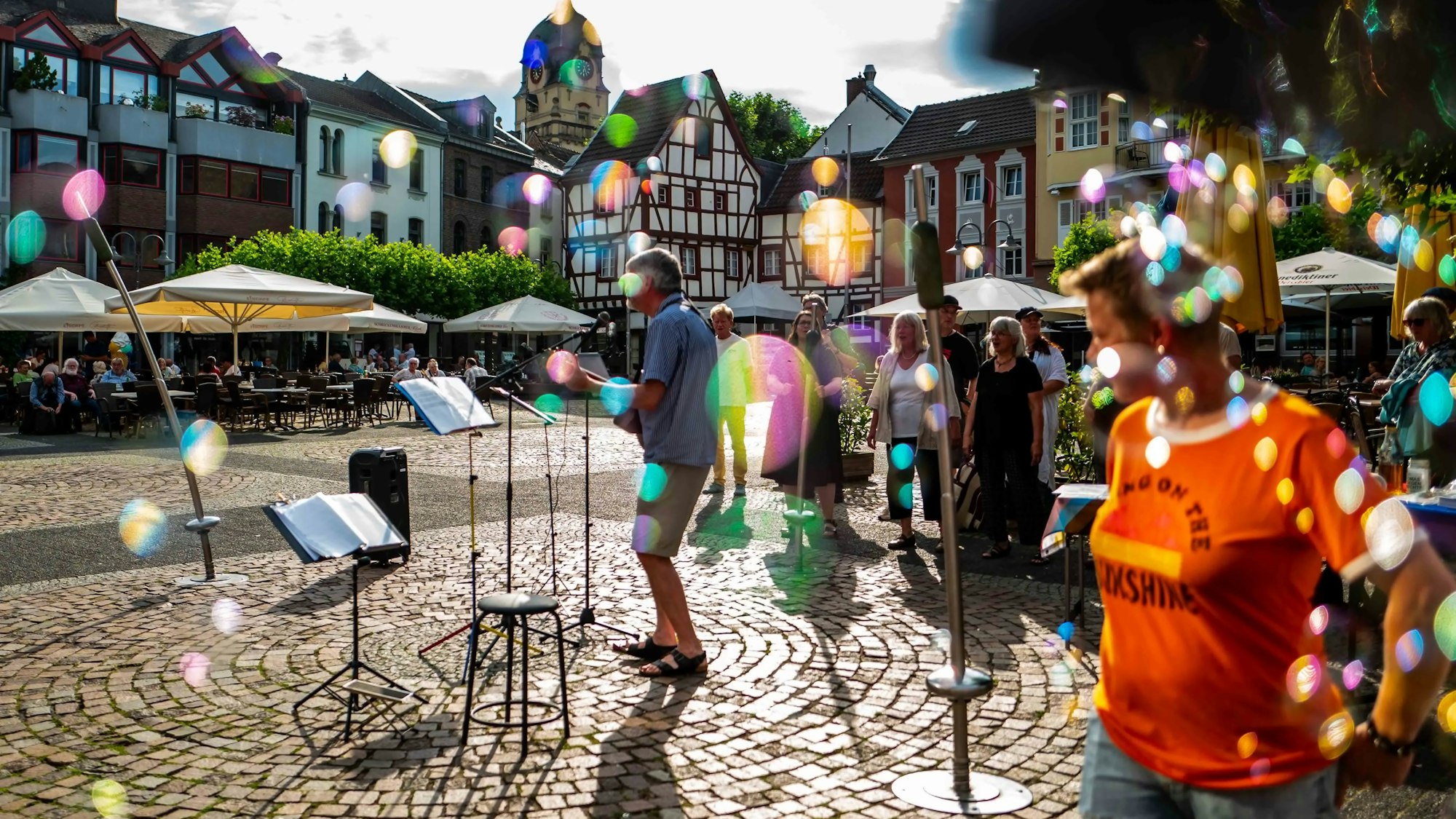 Das Bild zeigt einen Musiker auf dem Alten Markt in Euskirchen. Auf dem Bild sind zudem einige bunte Punkte zu sehen – das sind Seifenblasen im Sonnenlicht.