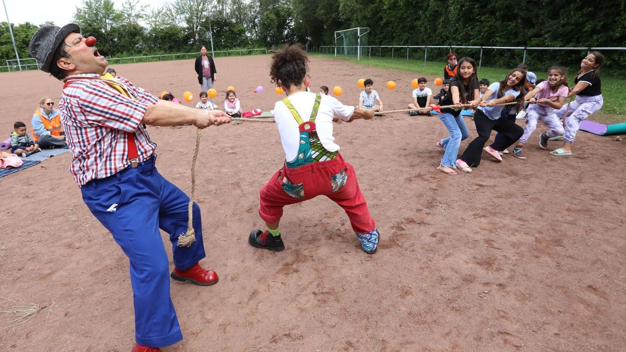 Zwei bunt gekleidete Clowns wetteifern beim Seil ziehen gegen eine Gruppe Kinder.