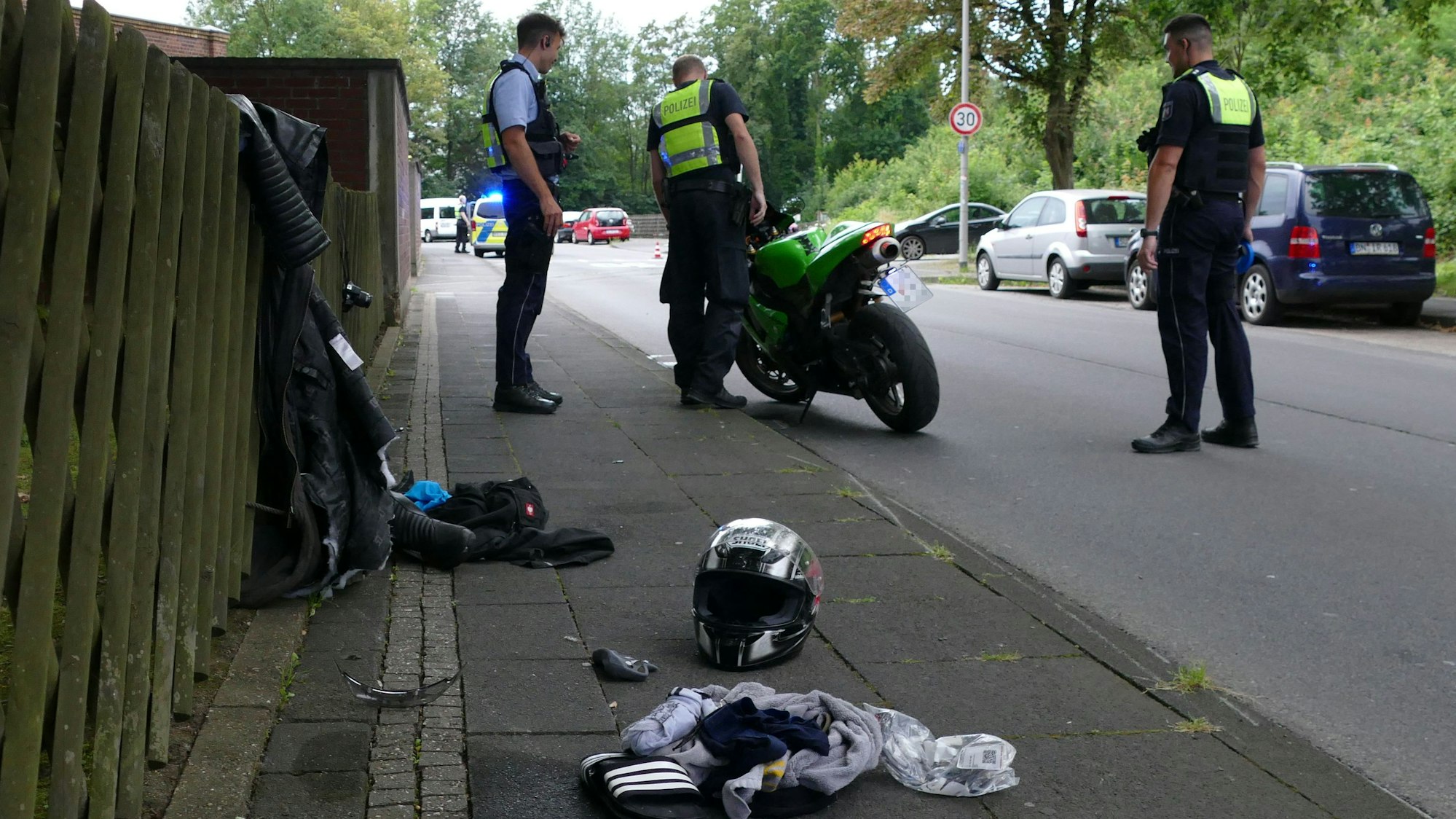 Polizisten in Dienstkleidung betrachten ein Motorrad. Auf dem Gehweg liegen Kleidungsstücke und ein Motorradhelm.
