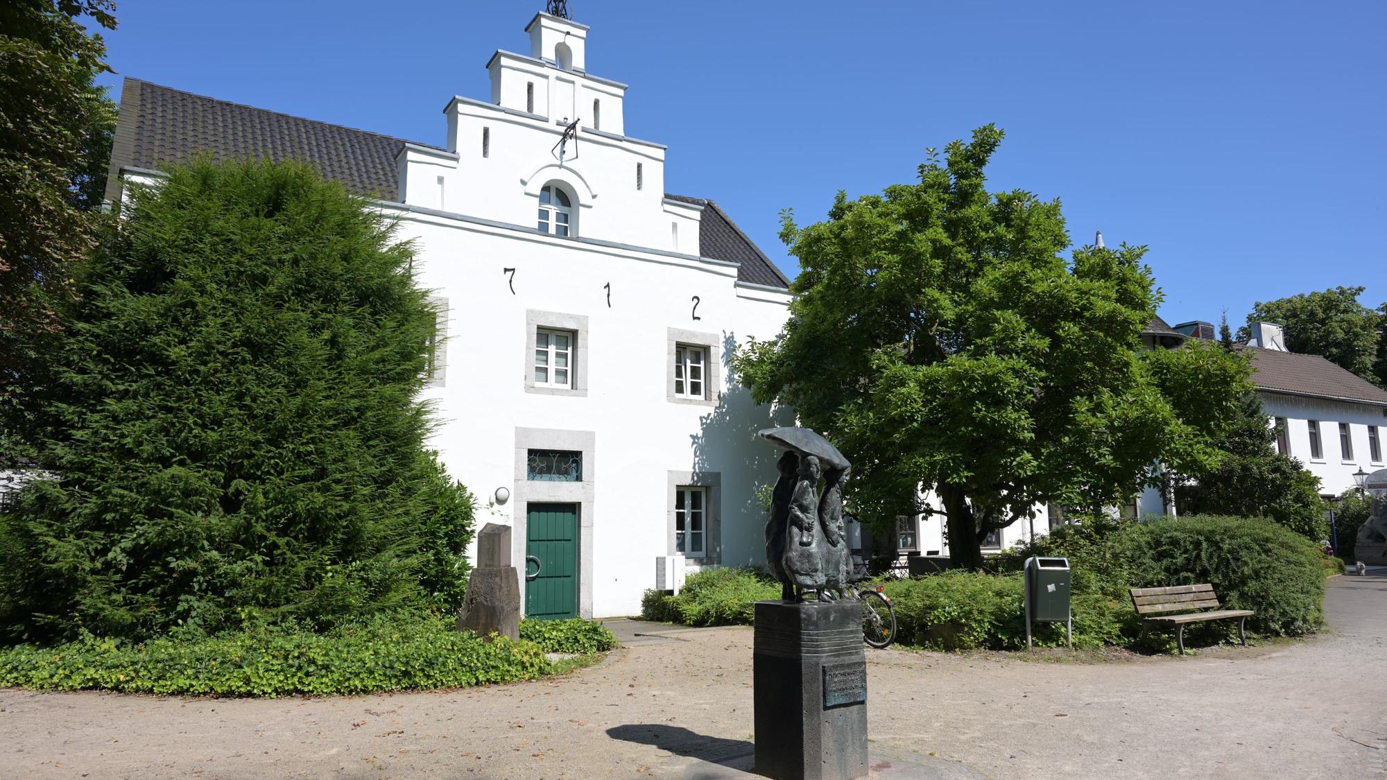 Das alte Gebäude mit weißem Anstrich ist von außen zu sehen. Davor steht ein Denkmal.