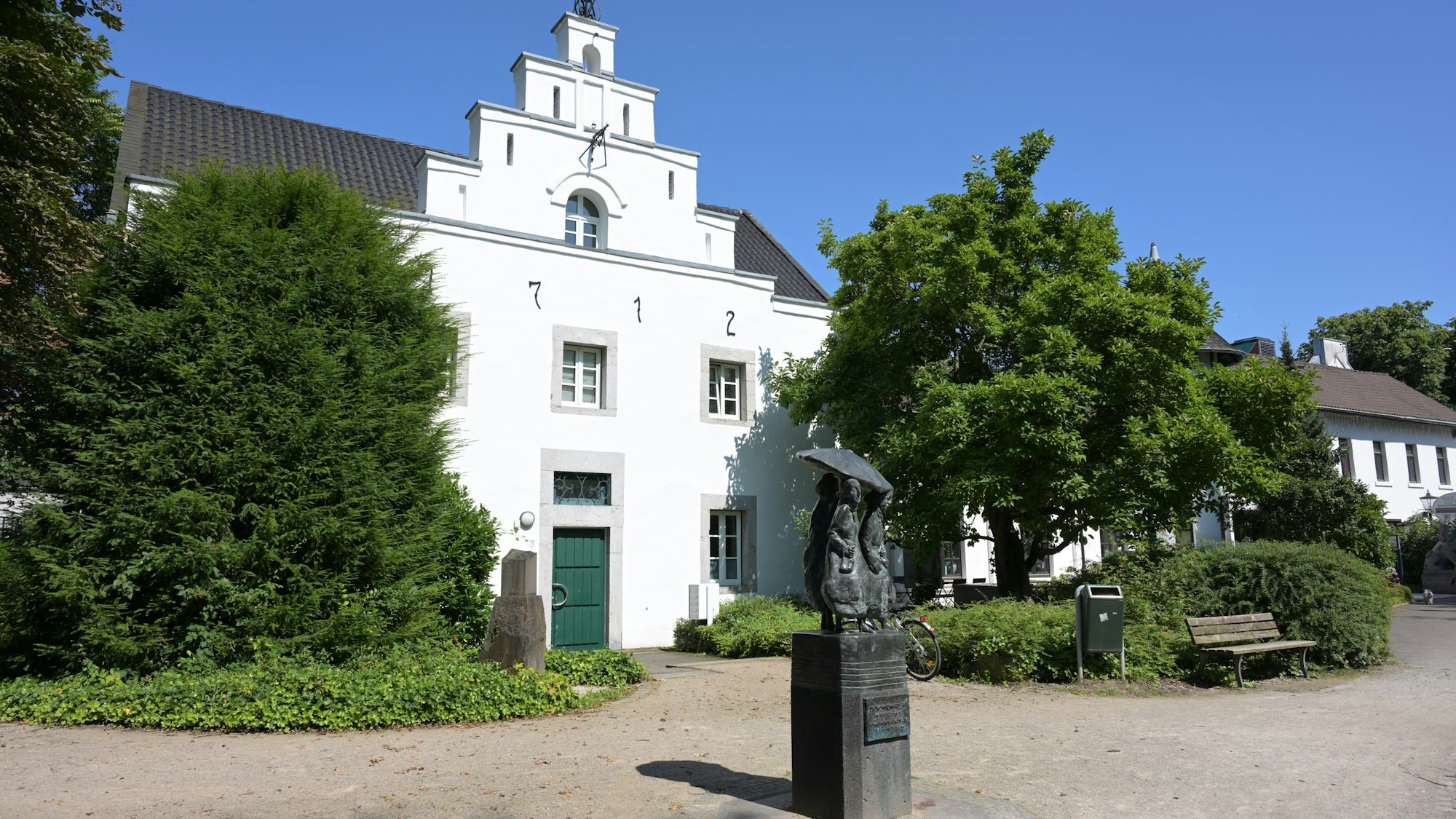 Das alte Gebäude mit weißem Anstrich ist von außen zu sehen. Davor steht ein Denkmal.