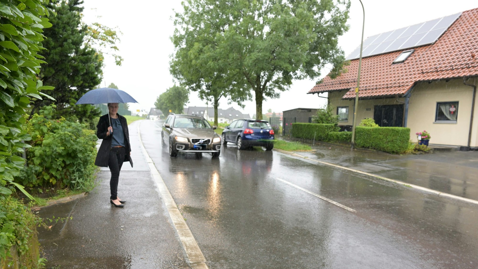 Eine Straße im Regen, zwei Autos fahren aneinander vorbei, eine Frau steht mit Regenschirm am, Straßenrand.