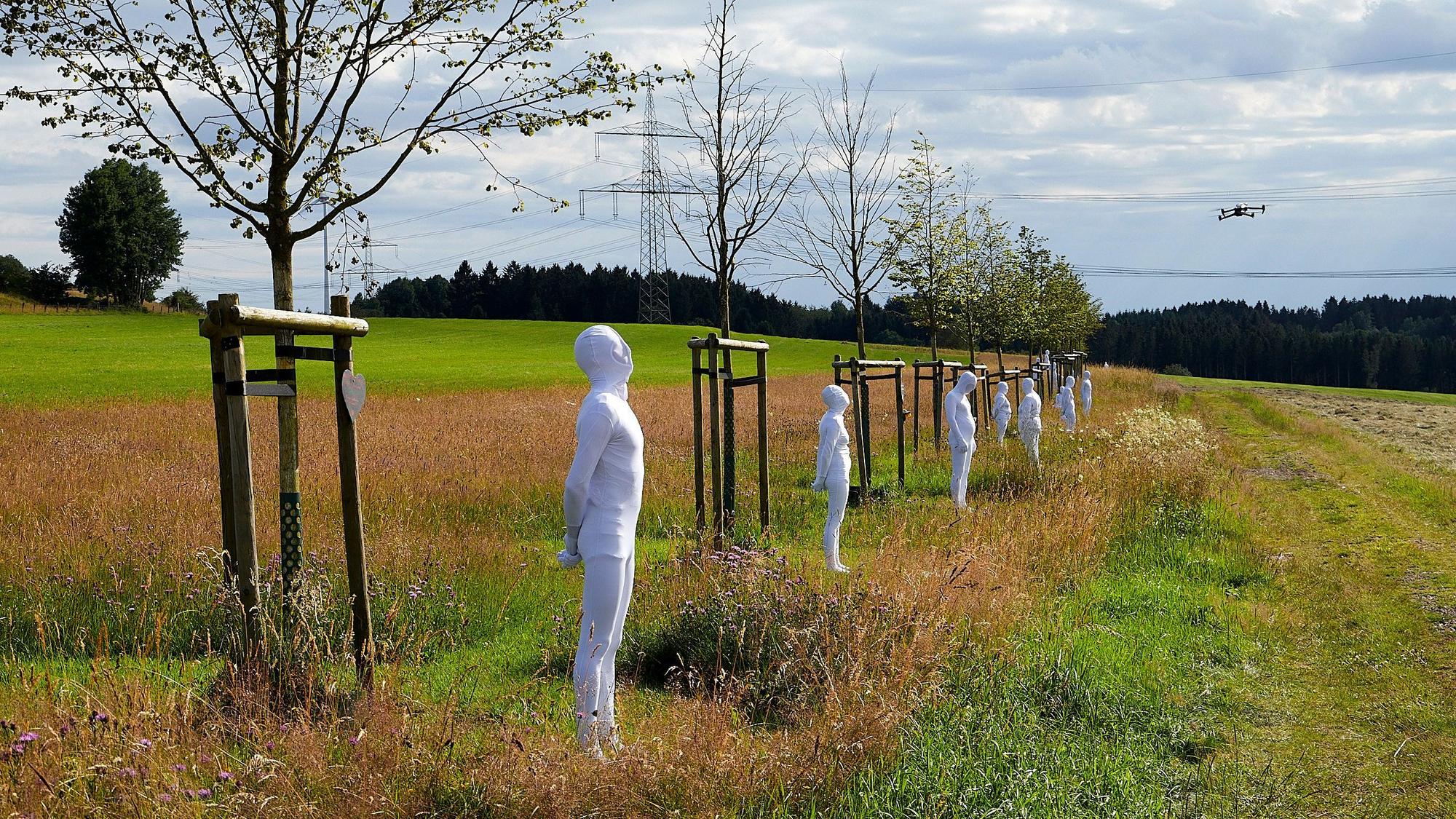 In weiße Anzüge gekleidete Menschen stehen vor den Bäumen der zentralen NRW-Gedenkstätte für die Opfer der Flutkatastrophe. Sie symbolisieren die gestorbenen Menschen.