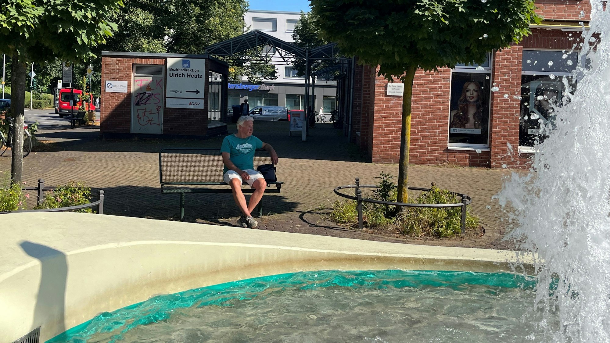 Rechts sieht man die Wasserfontäne. Der Kölner sitzt auf einer Bank.