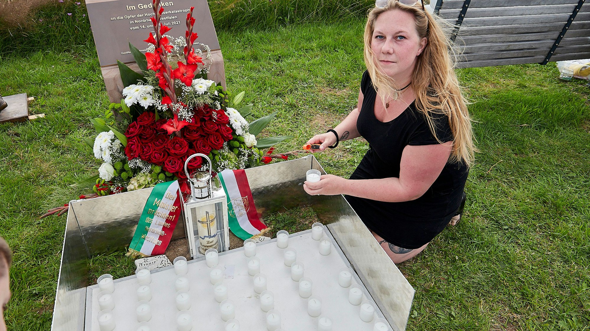 Tamara Kopelke kniet am Gedenkstein für die Opfer der Flutkatastrophe in Nordrhein-Westfalen, vor dem ein Blumengesteck steht. Vor der Frau ist eine Platte mit 49 Kerzen, die sie entzündet.