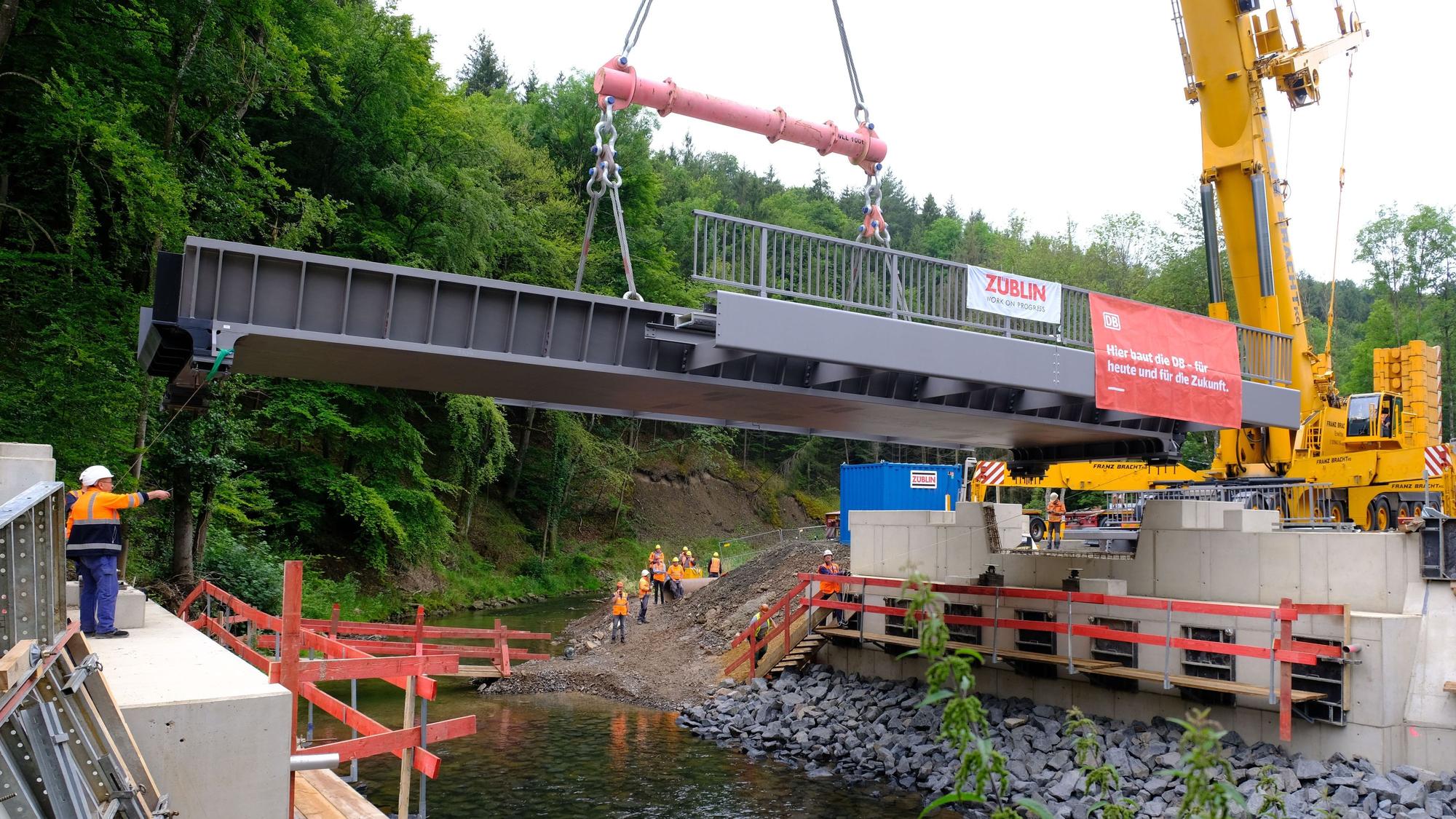 Mithilfe eines Mobilkrans wird eine Stahlbrücke auf die Widerlager gehoben. Darunter fließt die Urft in ihrem Flussbett.
