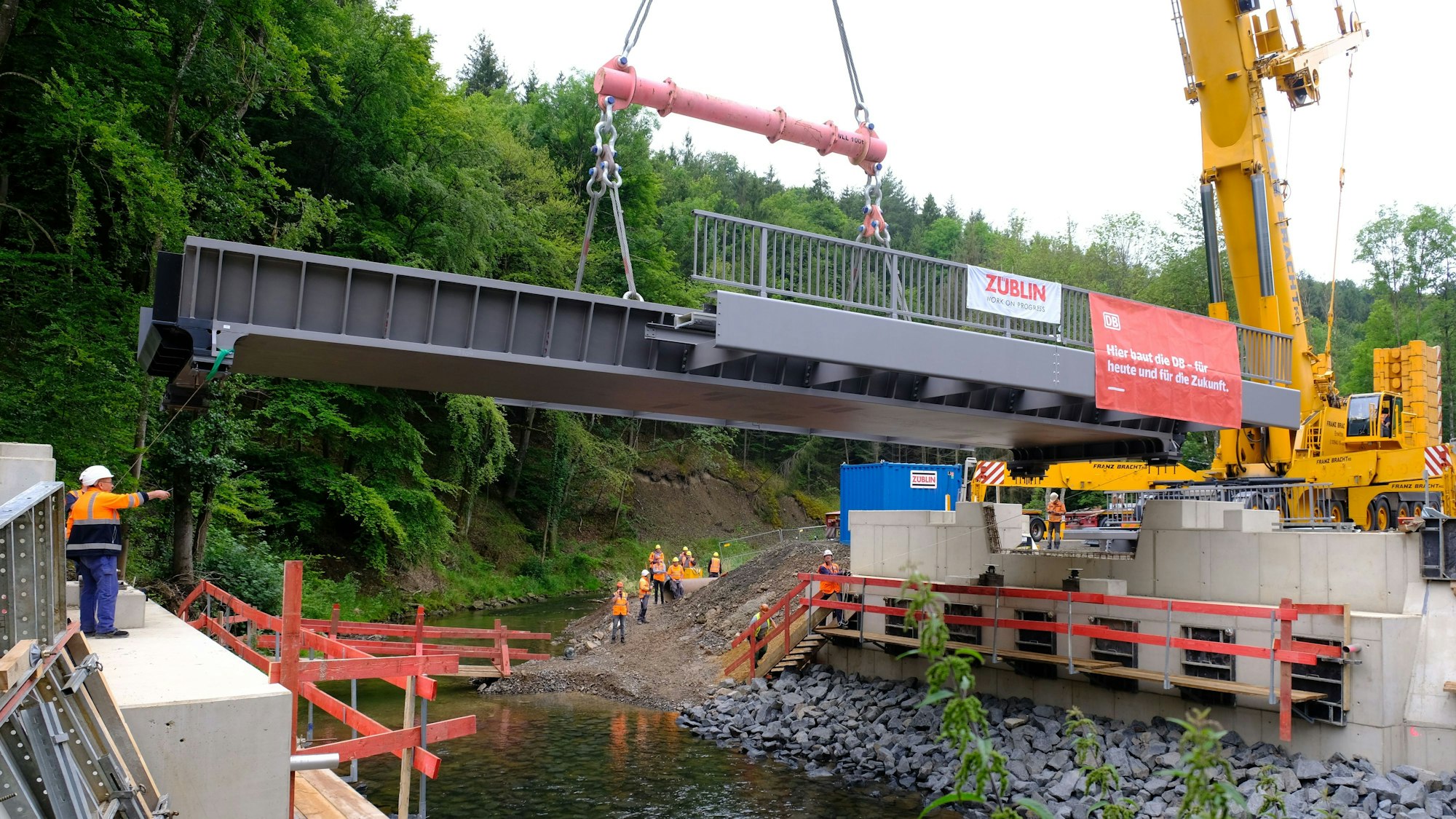 Mithilfe eines Mobilkrans wird eine Stahlbrücke auf die Widerlager gehoben. Darunter fließt die Urft in ihrem Flussbett.