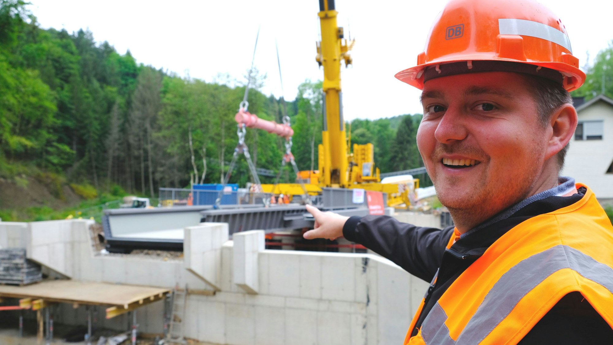 Projektleiter Felix Raffelsiefen trägt Schutzhelm und Warnweste. Er deutet auf die neue Brücke im Urfttal.