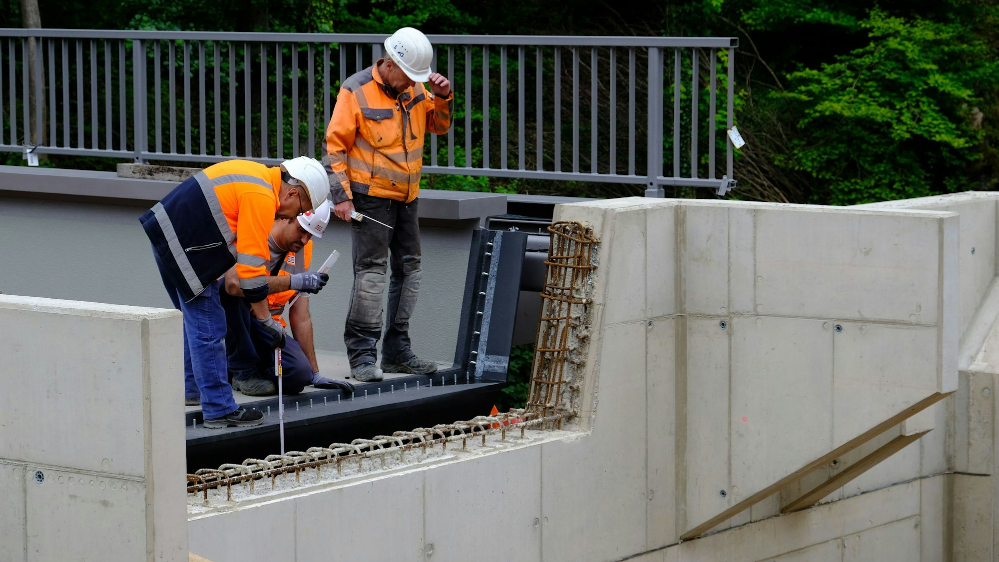 Drei Bauarbeiter messen mit Zollstöcken die Position einer neuen Brücke nach.