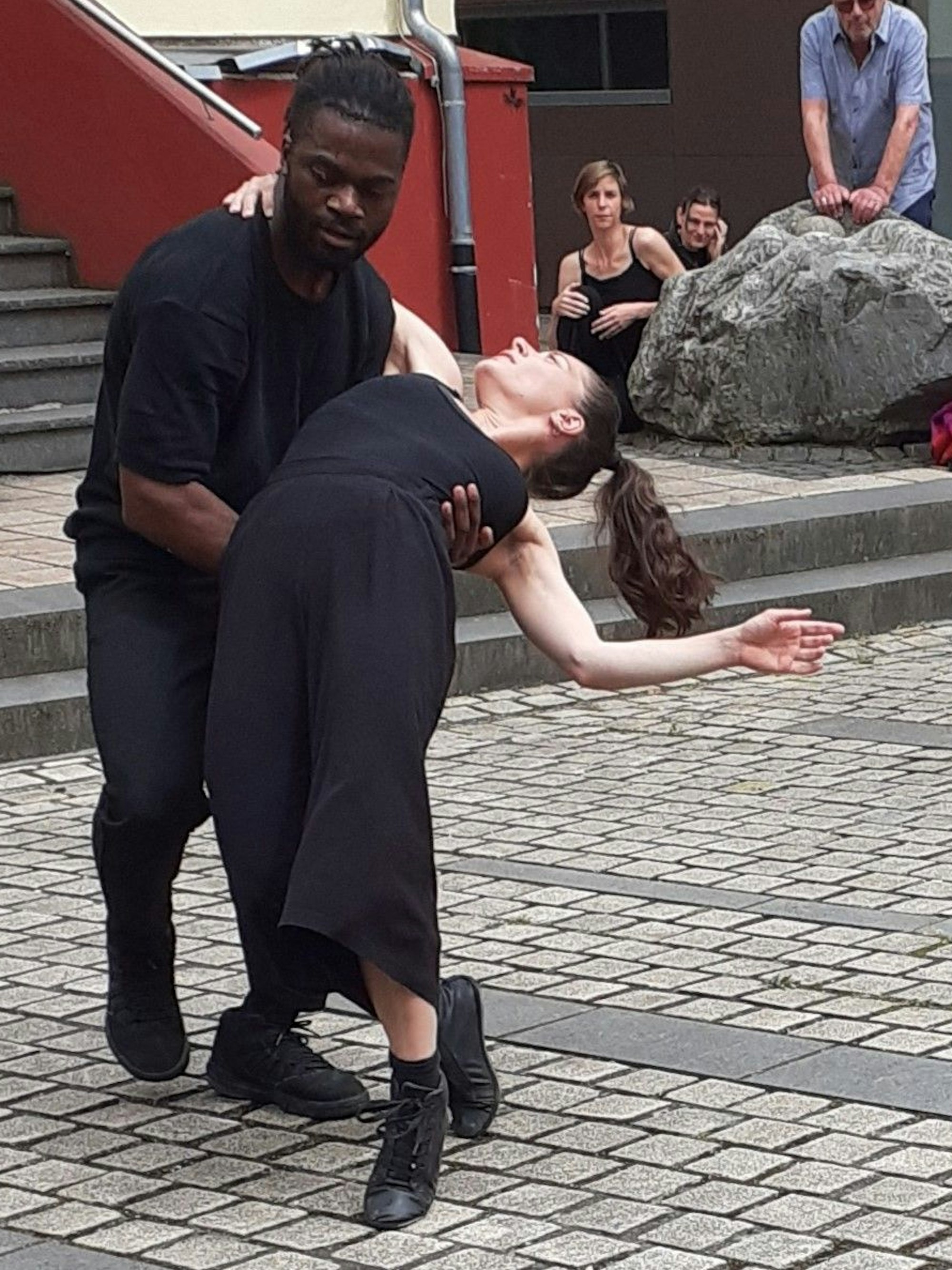 Eine Tänzerin und ein Tänzer bei ihrem Auftritt auf dem Platz vor dem Kunstforum Eifel. Er hat sie tänzerisch aufgefangen. Im Hintergrund sind ein paar Zuschauer und Akteure zu sehen.