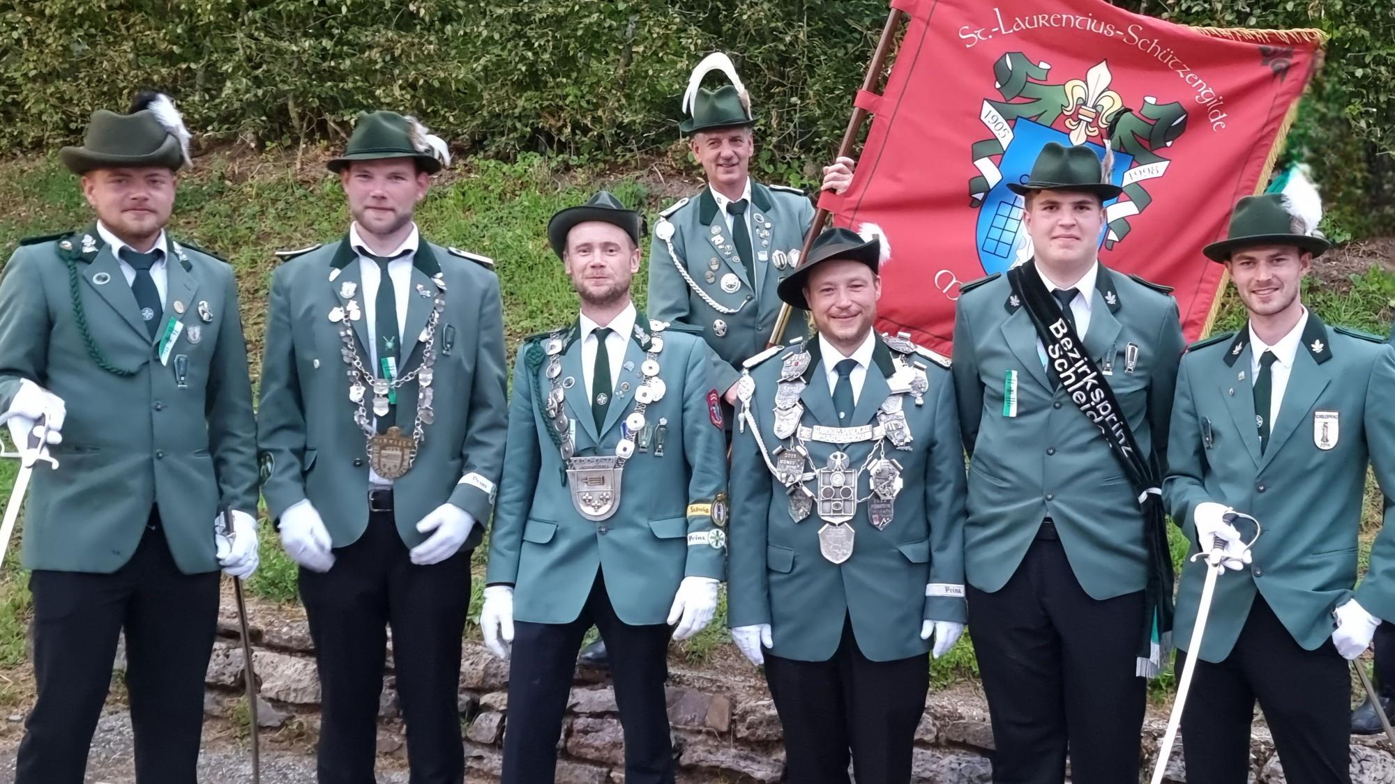 Sieben Männer in grünen Schützen-Uniformen stehen vor einer kleinen Mauer. Einer trägt die rote Fahne der Marmagener Gilde.