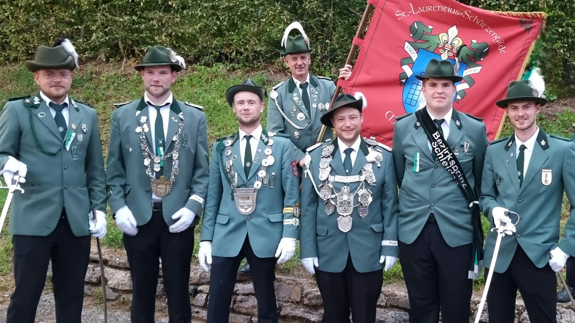 Sieben Männer in grünen Schützen-Uniformen stehen vor einer kleinen Mauer. Einer trägt die rote Fahne der Marmagener Gilde.