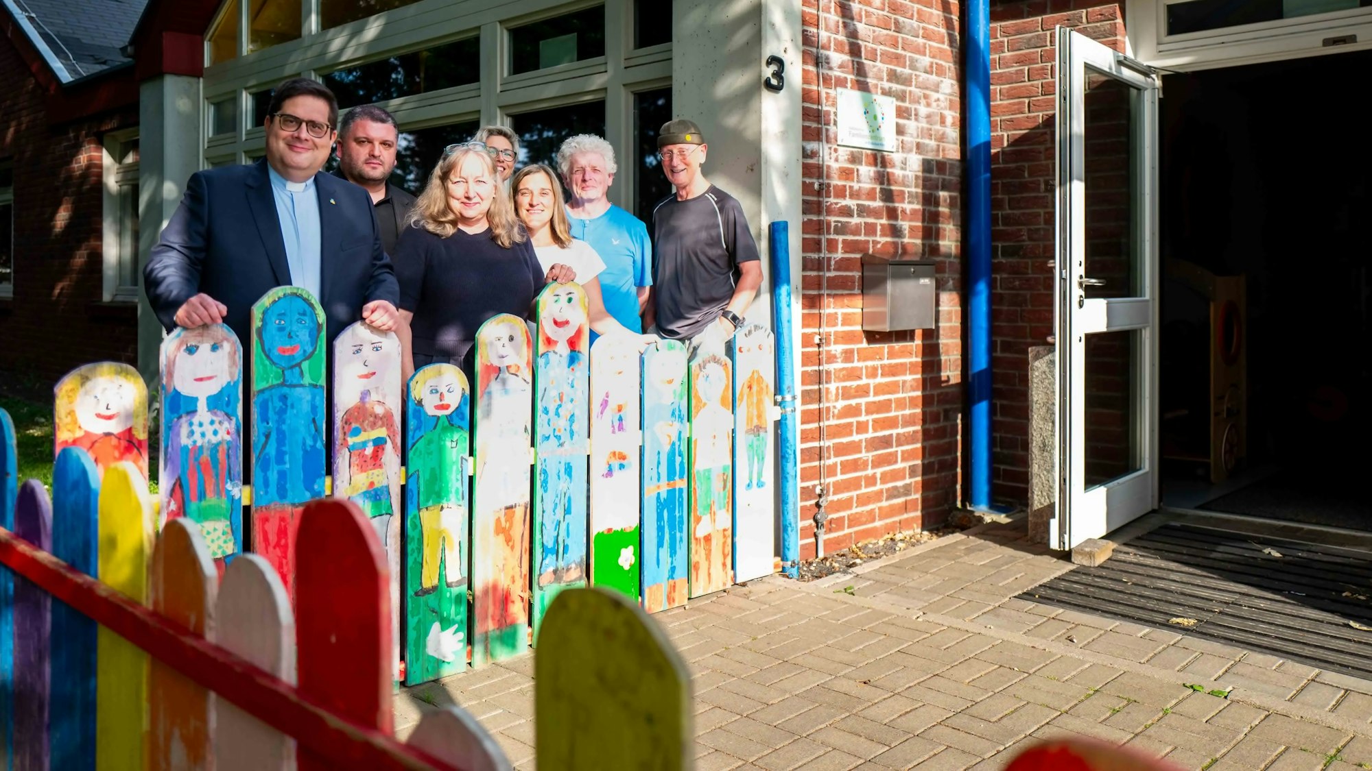 Das Bild zeigt einen Teil des Kirchenvorstands mit dem Leitenden Pfarrer vor der Kita in Kirchheim. Die Menschen stehen hinter einem Holzzaun, auf den Kindergesichter gemalt sind.