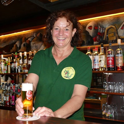 Frau in grünem T-Shirt steht hinter der Theke und stellt ein gefülltes Glas mit Kölsch auf den Tresen.