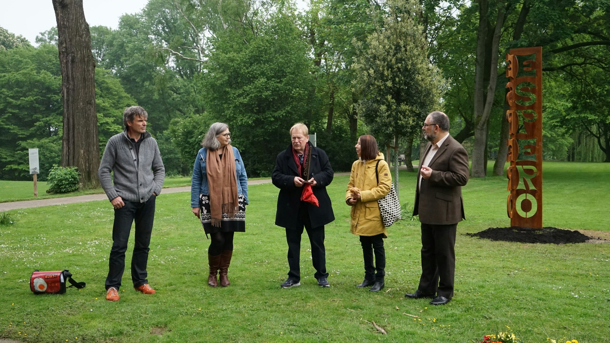 Drei Männer und zwei Frauen stehen in einem Park, rechts ist eine Stele zu sehen, gebildet aus dem Wort „Espero“, die Buchstaben stehen untereinander.