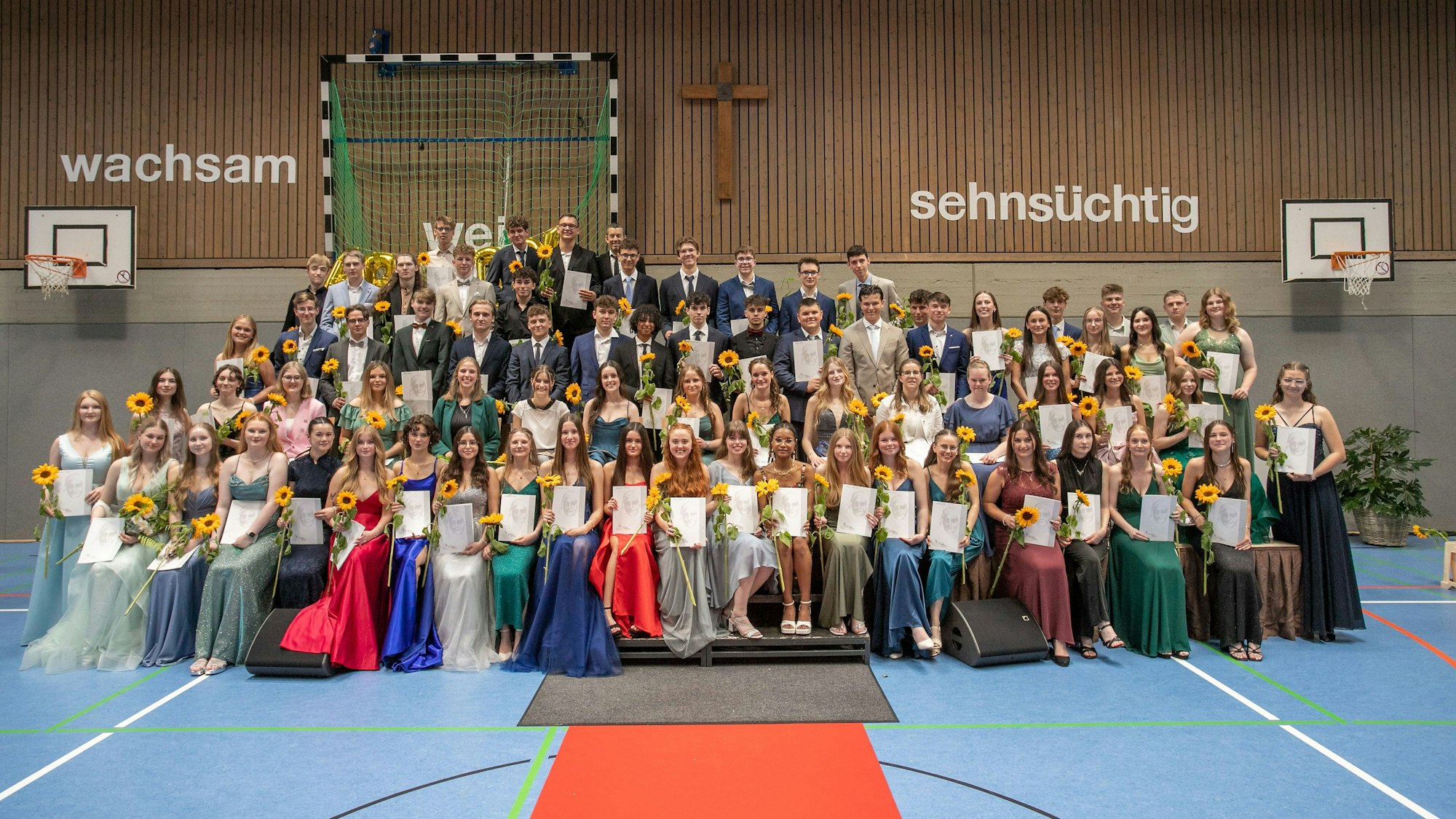 Die Abiturientia des St.-Angela Gymnasiums 2024 steht in Abendgarderobe und mit Sonnenblumen und Zeugnissen in der Hand in einer Turnhalle. An der Wand hängen die Worte: wachsam und sehnsüchtig.
