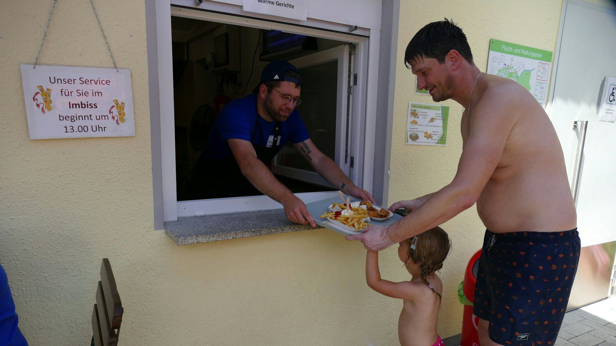 Ein Mann reicht ein Tablett mit Pommes und Currywurst aus dem Fenster eines Kiosks. Ein Kind und ein Mann in Badekleidung nehmen das Tablett an.