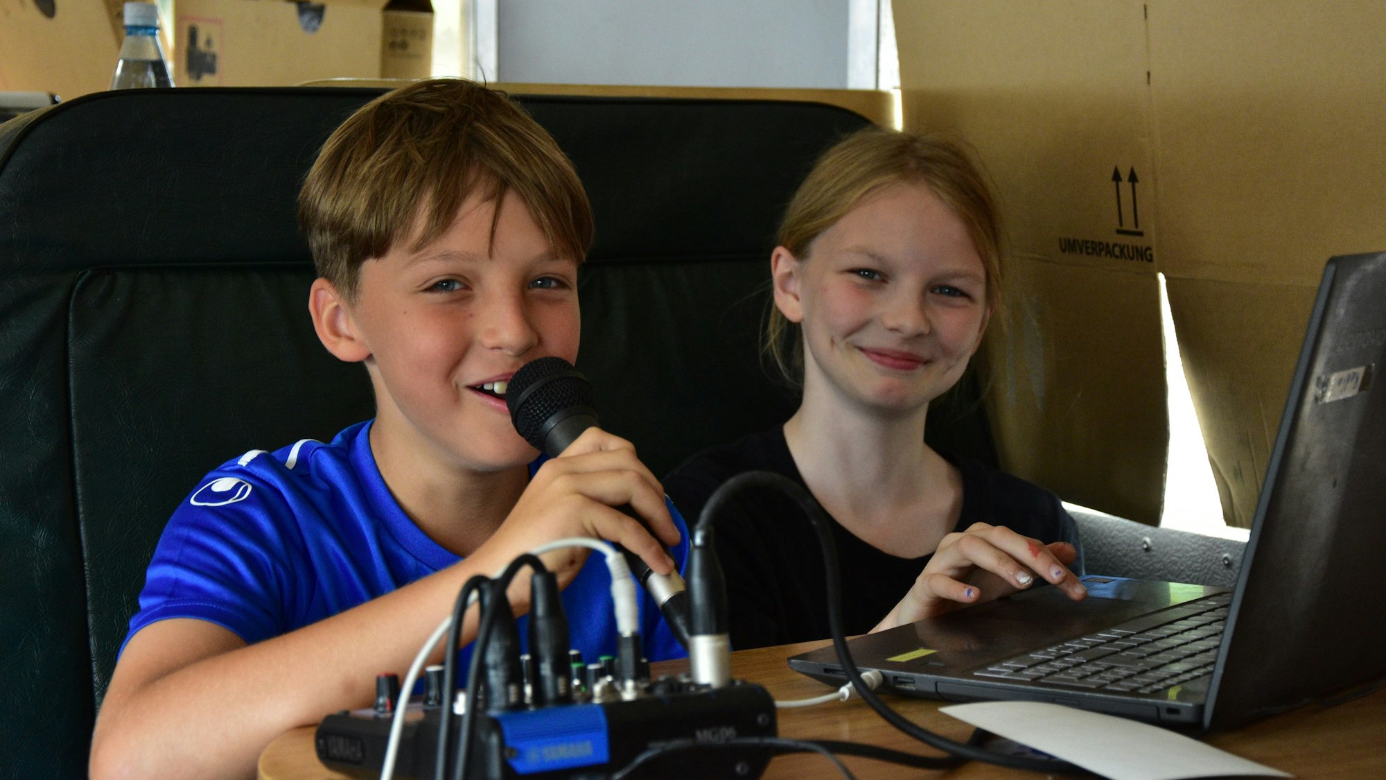 Ein Junge und ein Mädchen an einem Mischpult mit Computer. Der Junge hat ein Mikrophon in der Hand.