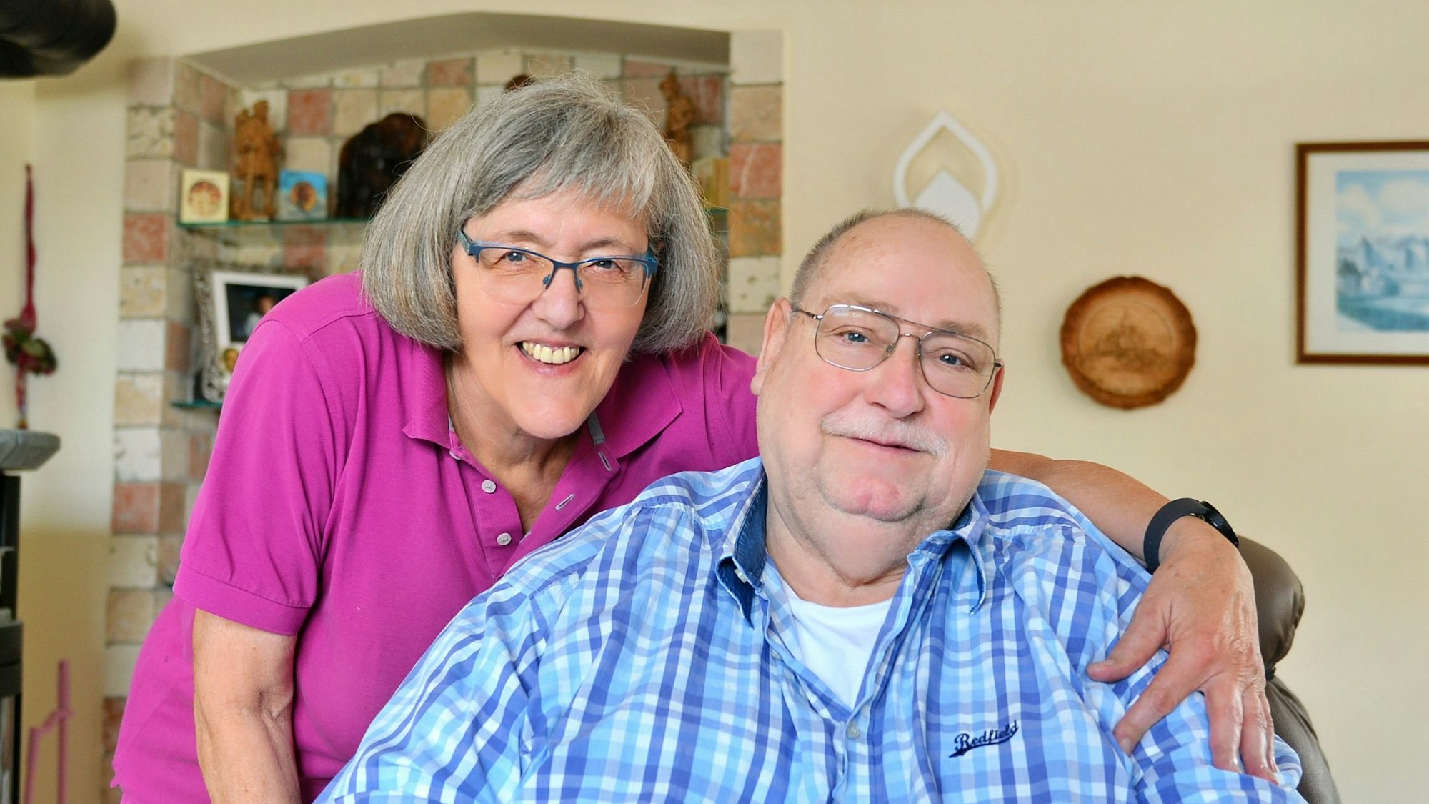 Die Eheleute schauen freundlich in die Kamera. Karin Benden umarmt ihren Mann, der auf einem Sessel sitzt, während sie neben ihm steht.