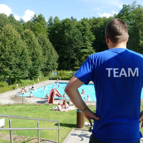 Ein Mann schaut auf ein Schwimmbad. Er trägt ein blaues T-Shirt, auf dem Rücken steht Team.