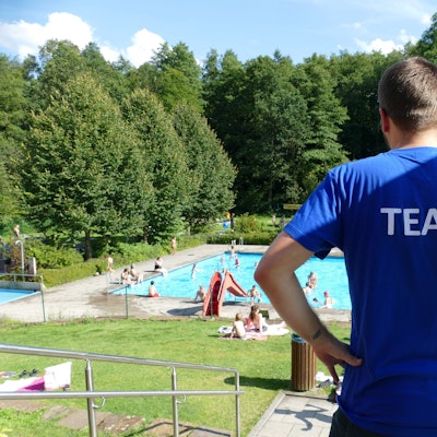 Ein Mann schaut auf ein Schwimmbad. Er trägt ein blaues T-Shirt, auf dem Rücken steht Team.