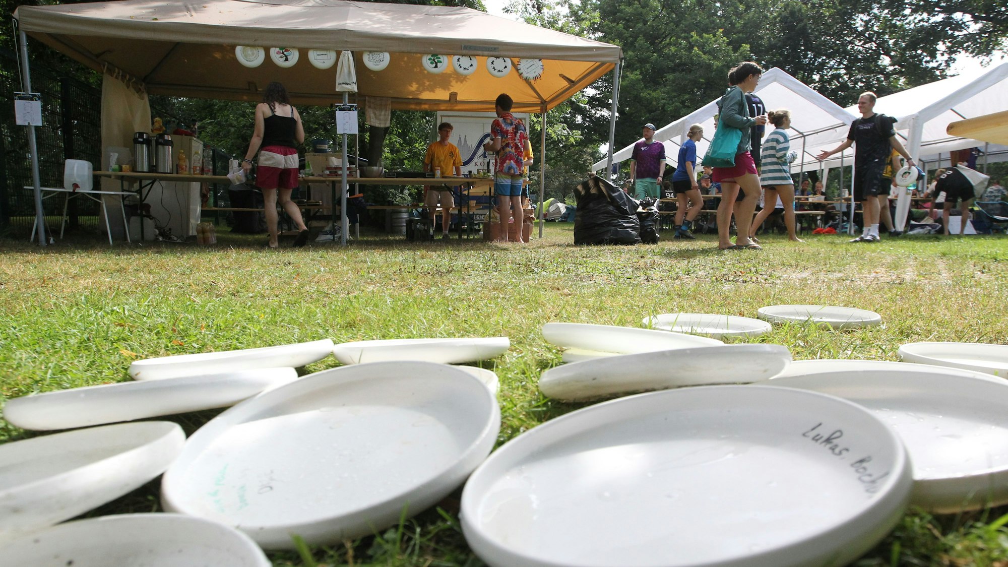 Das Bild zeigt im Vordergrund ein Dutzend herumliegender, weißer Frisbees und auf der Wiese dahinter mehrere Pavillonzelte.