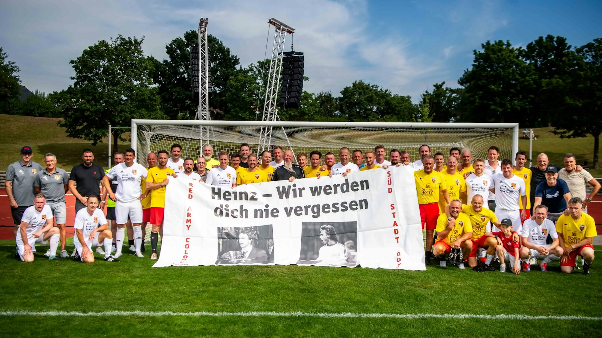 Zwei Fußballmannschaften in gelben und weißen Trikots stehen gemeinsam vor einem Tor. Sie halten ein großes Transparent mit zwei Fotos von Heinz Flohe und der Aufschrift „Heinz - wir werden dich nie vergessen“.
