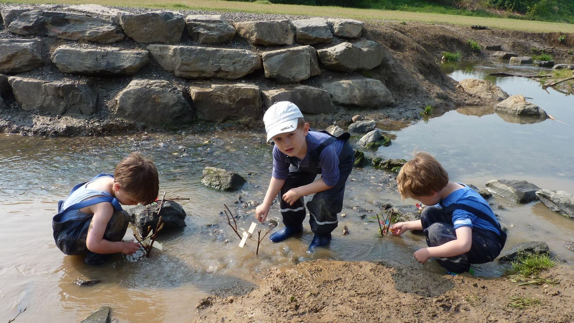 Drei Kitakinder hocken im Wasser, sie testen, ob ihre Wassermühlen funktionieren.
