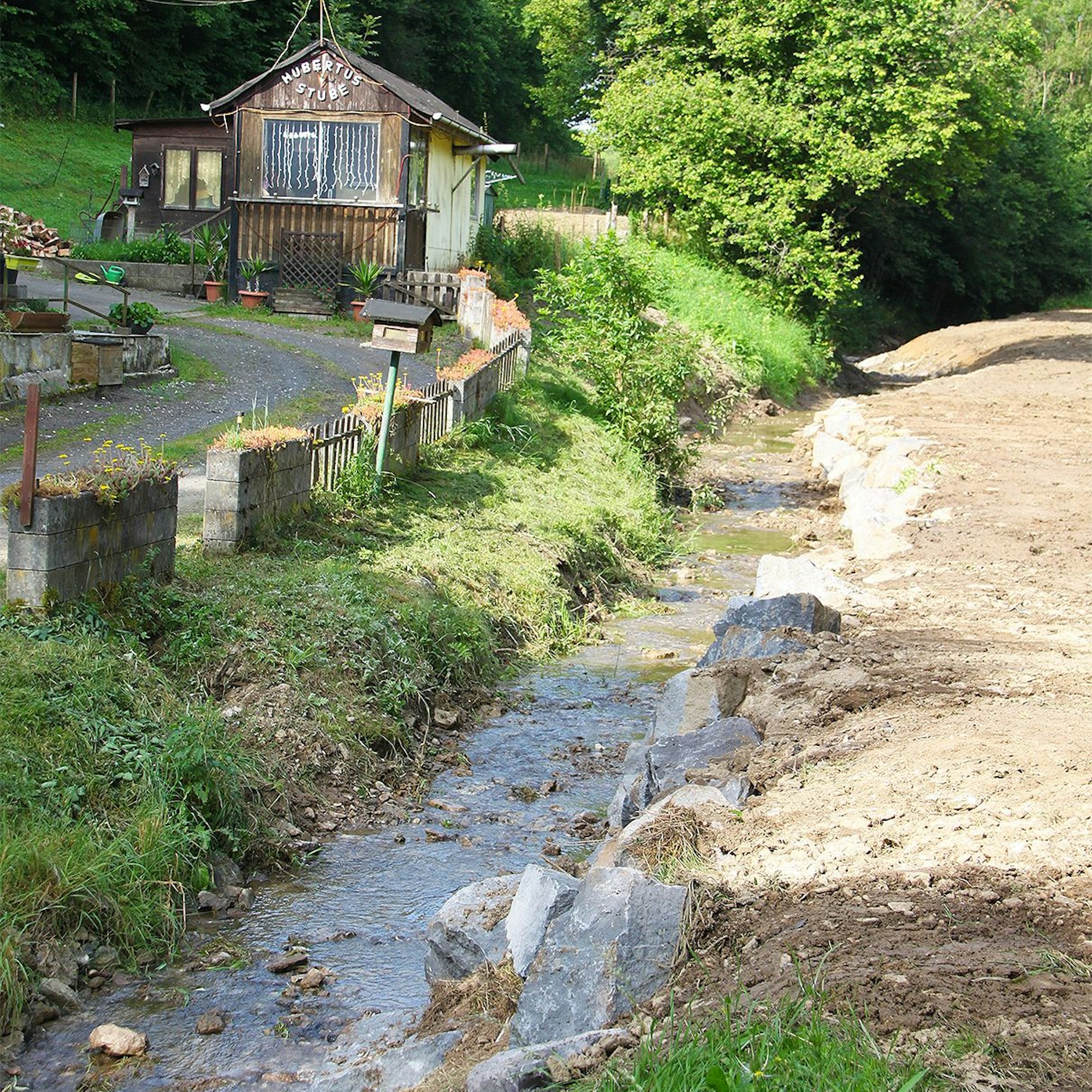 Das Bild zeigt den Fuhrbach in der Gemeinde Blankenheim kurz vor der Mündung in die Ahr. Im Hintergrund ist ein kleines Haus, auf dessen Giebel in weißer Schrift „Hubertusstube“ steht.