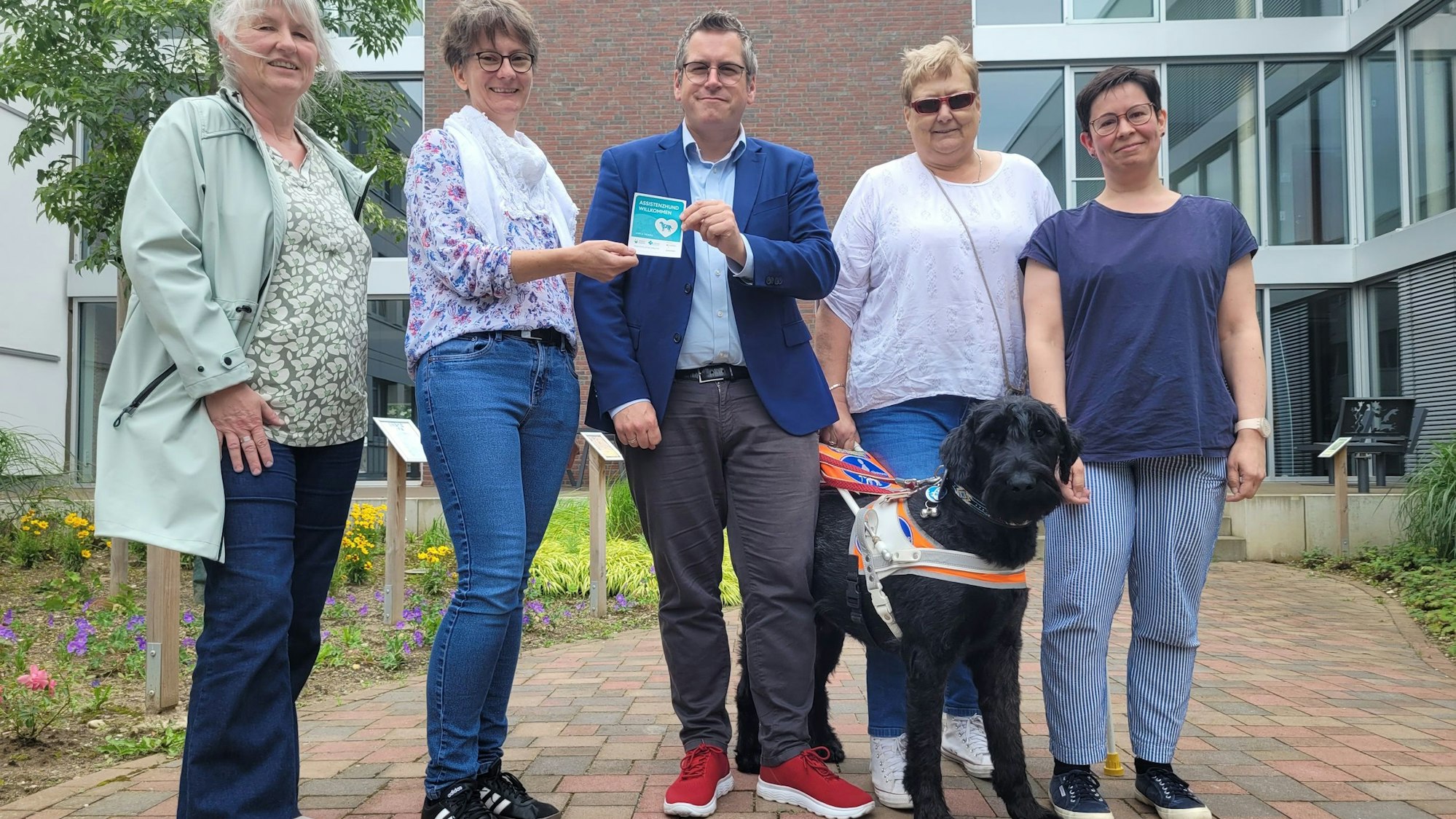 Vier Frauen und ein Mann lächeln in die Kamera, zwei Personen halten einen Aufkleber mit der Aufschrift „Assistenzhund willkommen“ in der Hand. Darum ist auch ein Hund dabei.