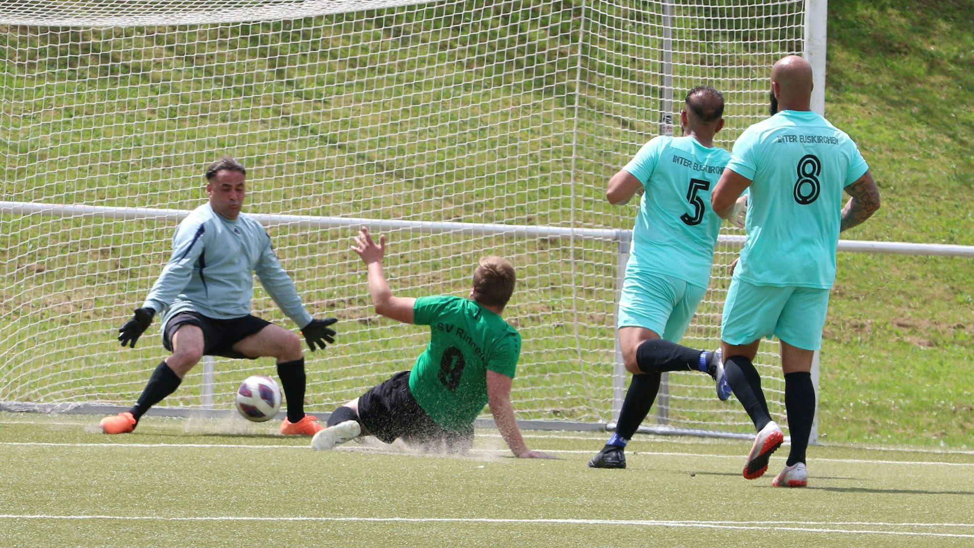 Ein Stürmer schiebt liegend den Ball zum Tor ein. Dabei bleibt der Torwart offenbar im Rasen hängen und verletzt sich.
