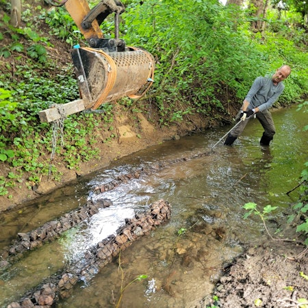 Ein Mann zieht mit einem Haken ein längliches, mit kleinen Steinen gefülltes Gebilde, an die richtige Position in einem Bachbett.