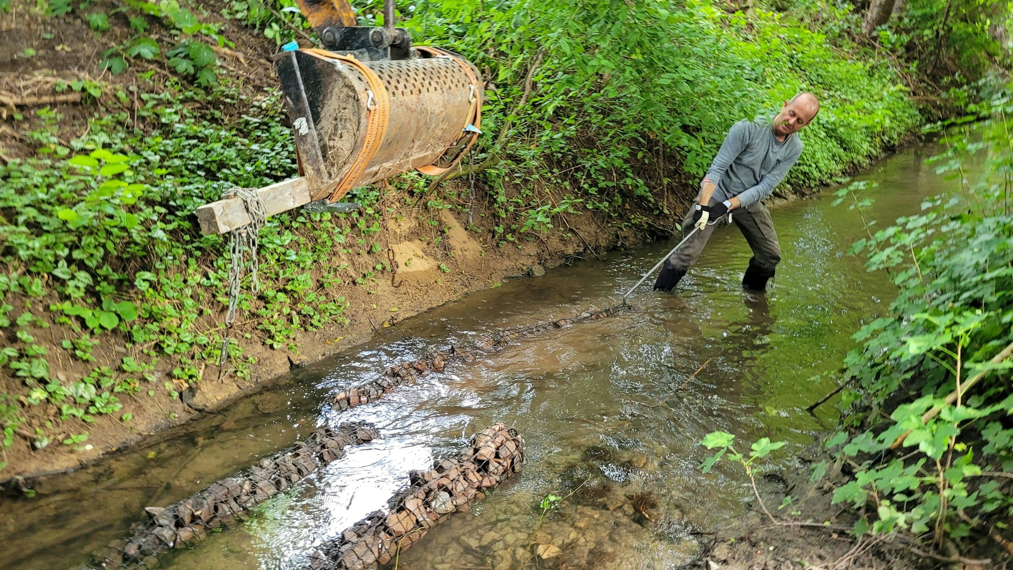 Ein Mann zieht mit einem Haken ein längliches, mit kleinen Steinen gefülltes Gebilde, an die richtige Position in einem Bachbett.