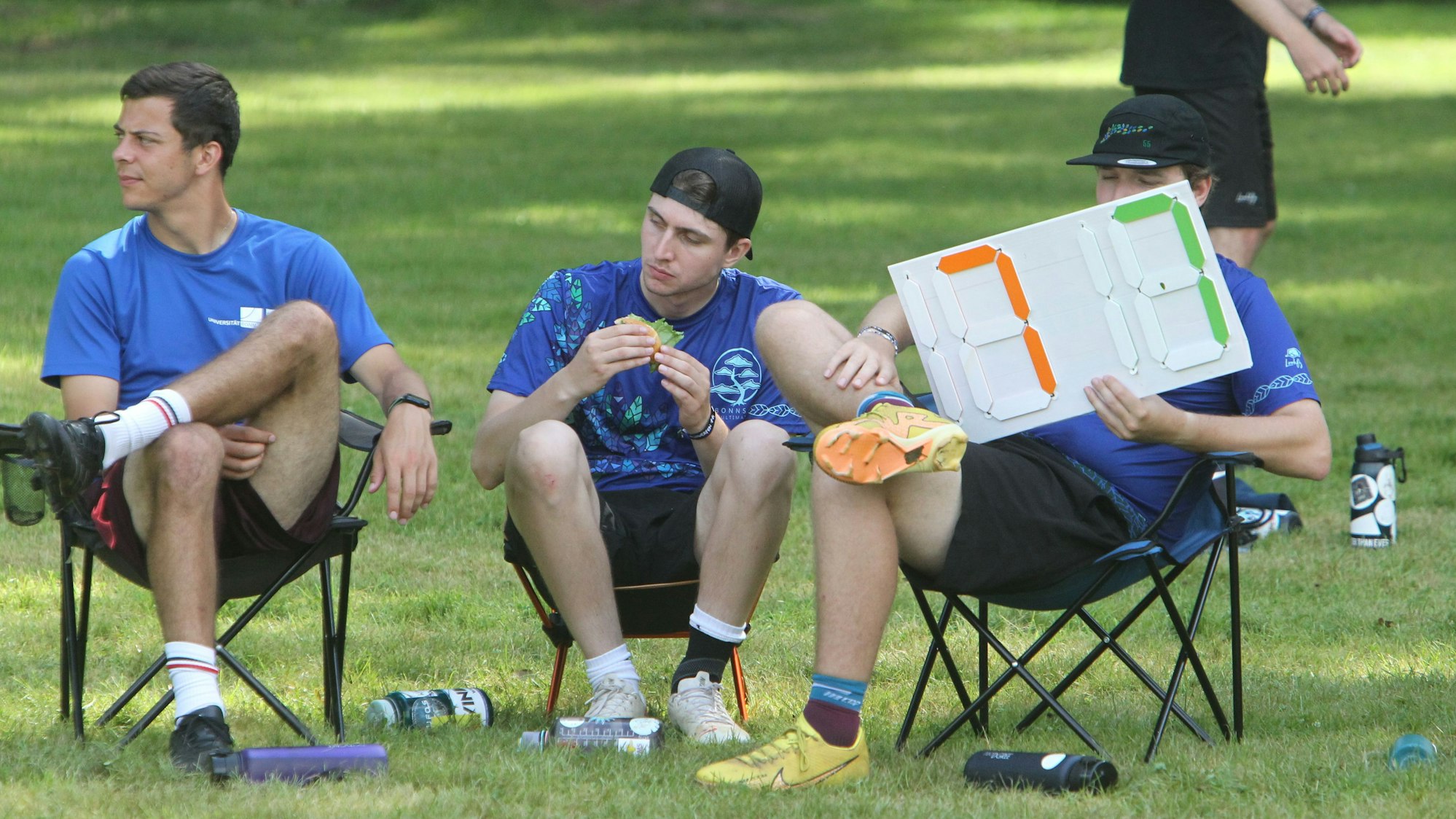 Drei junge Männer sitzen in Campingstühlen am Spielfeldrand. Einer zeigt mit einer Tafel den Spielstand.