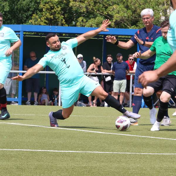 Vollen Körpereinsatz zeigt ein Fußballer, der mit ausgestreckten Armen und Ausfallschritt vor dem Gegenspieler an den Ball kommt. Der Schiedsrichter, Mitspieler und Zuschauer haben die Situation im Blick.