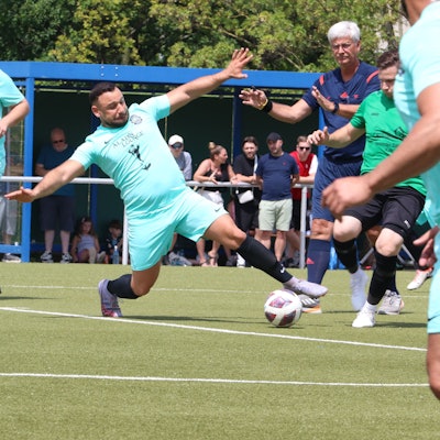 Vollen Körpereinsatz zeigt ein Fußballer, der mit ausgestreckten Armen und Ausfallschritt vor dem Gegenspieler an den Ball kommt. Der Schiedsrichter, Mitspieler und Zuschauer haben die Situation im Blick.