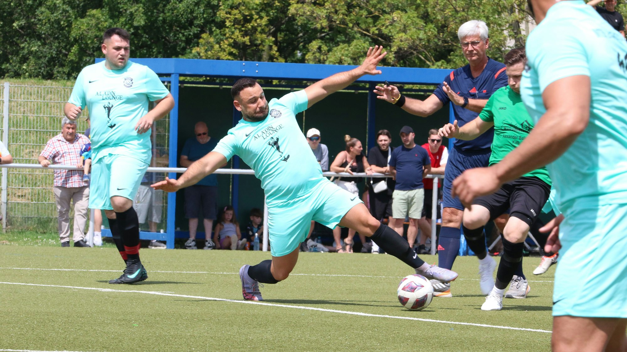 Vollen Körpereinsatz zeigt ein Fußballer, der mit ausgestreckten Armen und Ausfallschritt vor dem Gegenspieler an den Ball kommt. Der Schiedsrichter, Mitspieler und Zuschauer haben die Situation im Blick.