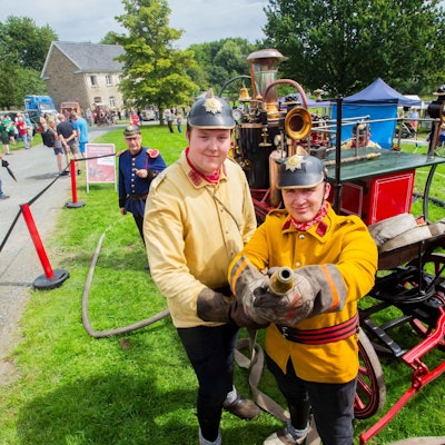 Zwei Männer in historischen Feuerwehr-Uniformen zielen mit einem Strahlrohr in die Kamera, hinter ihnen ist eine Dampfspritze aus dem Jahr 1901 zu erkennen.